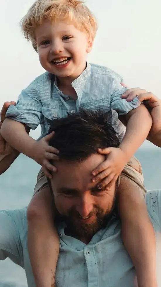 Smiling young boy riding piggyback on his father's shoulders, with the father smiling while holding his son's arms outdoors