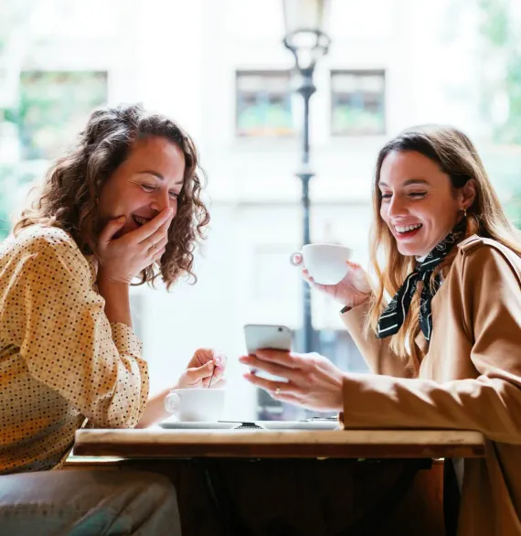 Two women laughing together while looking at a phone over coffee.