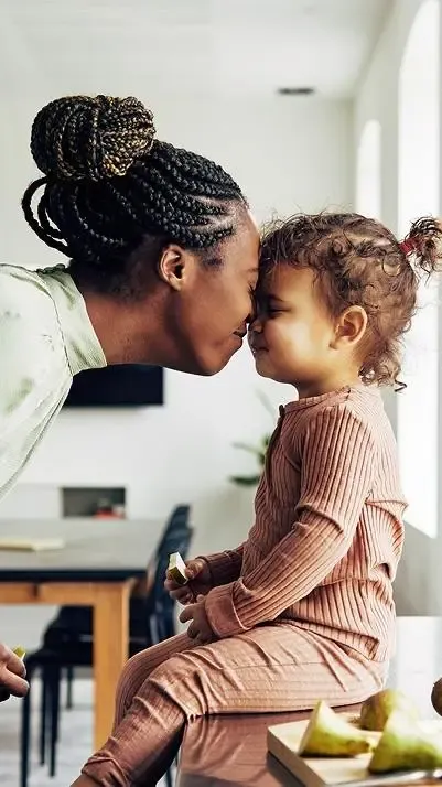A mother leaning forward to gently touch foreheads with her toddler daughter, who is sitting on a counter near sliced pears