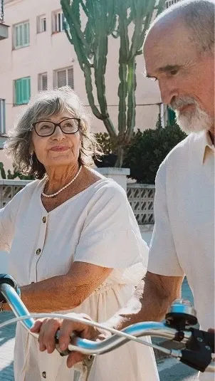 An older woman wearing glasses and a pearl necklace looking affectionately at her partner while standing together with bicycles outdoors