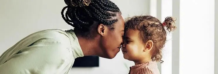 A mother leaning forward to gently touch foreheads with her young daughter indoors