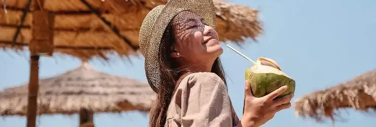 Woman in a straw hat sipping from a fresh coconut under a thatched umbrella at the beach