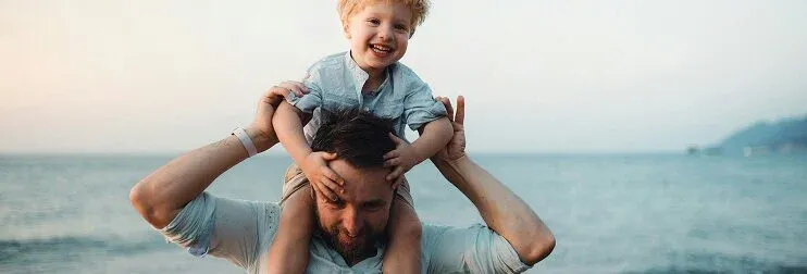 A laughing young boy sitting on his father's shoulders at the beach near the water at sunset