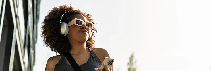 A woman with curly hair, sunglasses, and over-ear headphones holding a smartphone outdoors