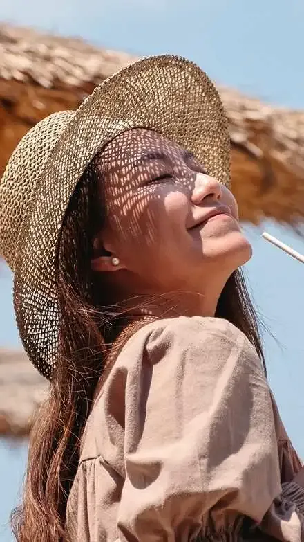 Woman wearing a woven straw hat tilting her head back to catch the sunlight while holding a drink