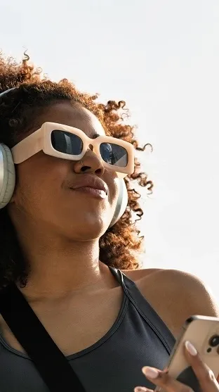 Woman with curly hair wearing white-framed sunglasses and over-ear headphones while holding a smartphone