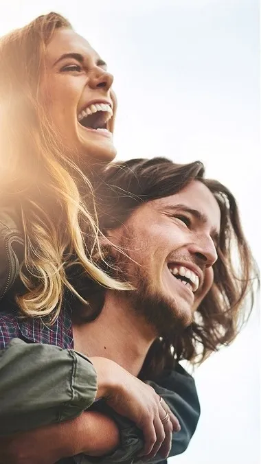 Close-up of a smiling man with shoulder-length hair giving a piggyback ride to a laughing woman outdoors